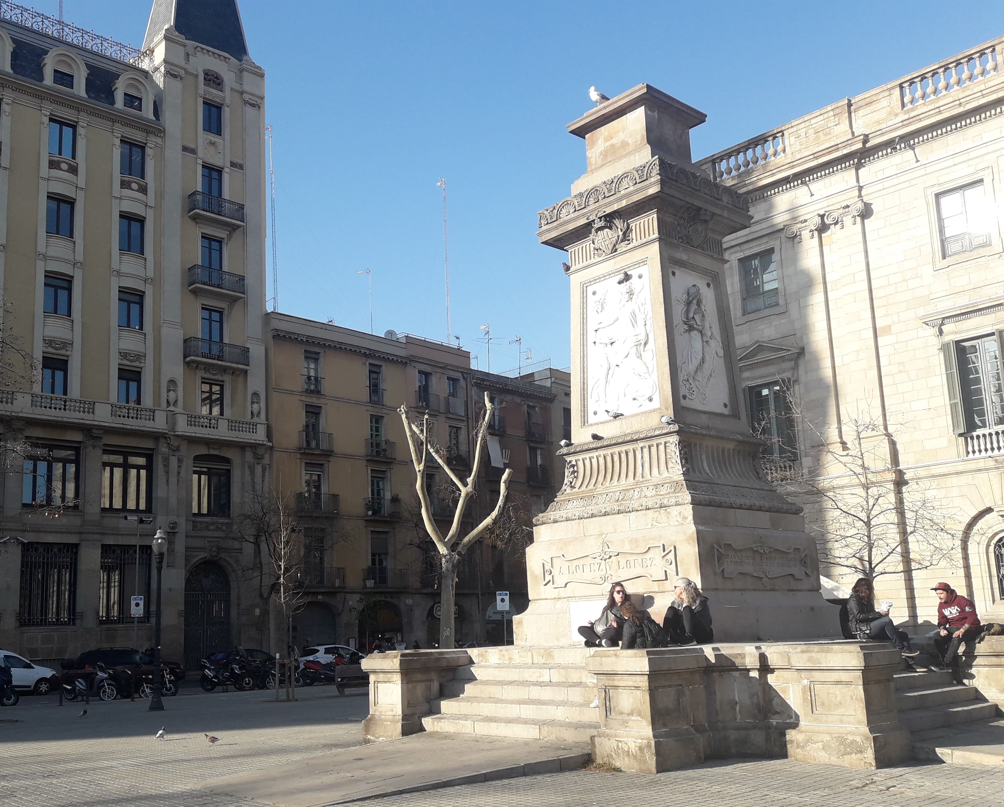 Pedestal del monumento al traficante de africanos Antonio López en Barcelona, con inscripciones explicativas de facto ilocalizables delante de los coches, al fondo a la izquierda